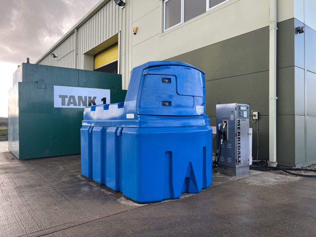 An outdoor fleet refueling station featuring a large blue plastic AdBlue storage tank positioned next to a stainless steel Merridale fuel dispensing pump. In the background, a large green metal fuel tank labeled "TANK 1" sits adjacent to a commercial commercial building.