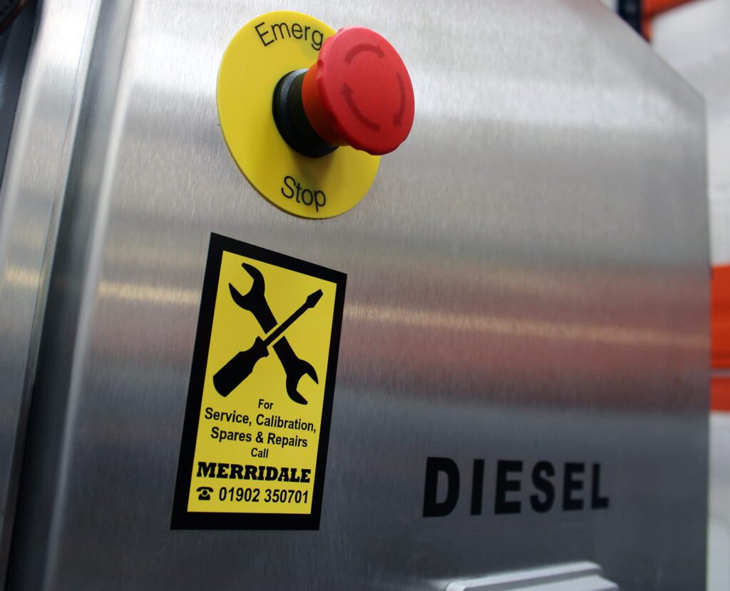 A close-up of a stainless steel industrial diesel pump featuring a large red emergency stop button and a yellow service sticker. The sticker displays a crossed wrench and screwdriver icon with contact information for Merridale service, calibration, spares, and repairs.