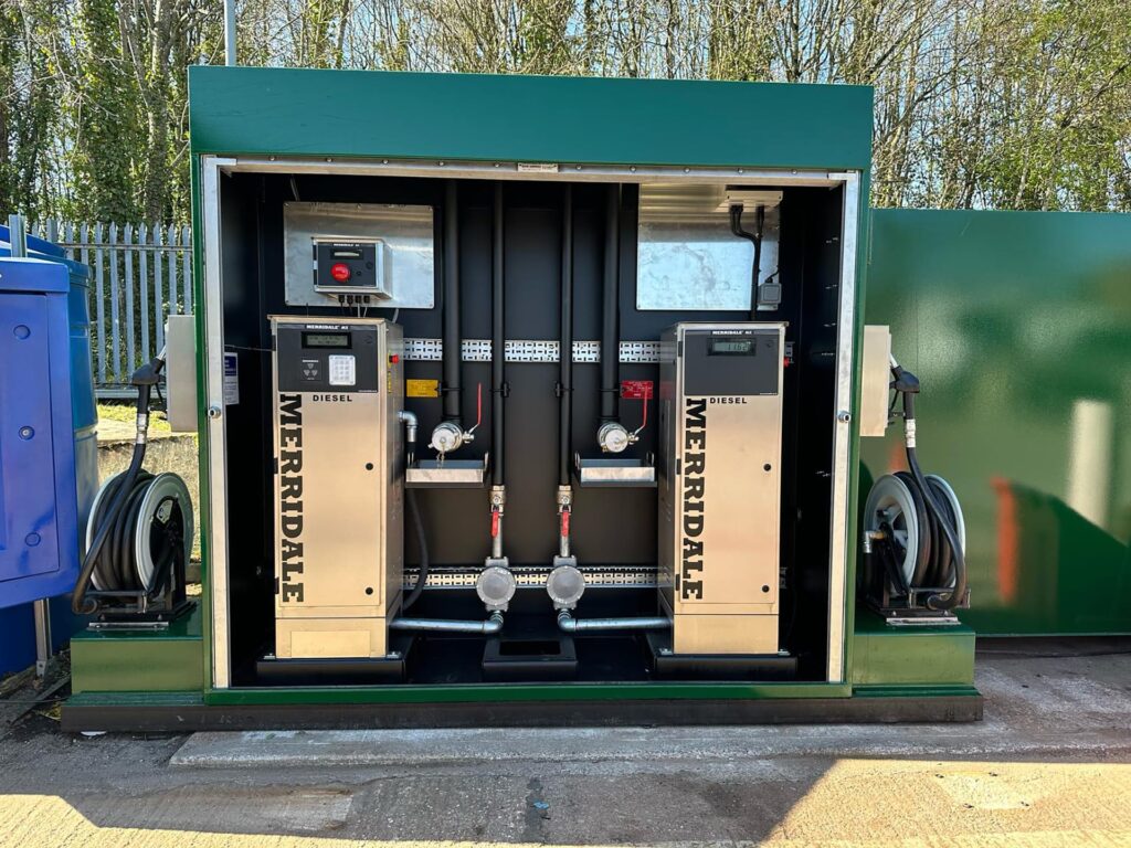 A symmetric, front-facing view of a dual-pump Merridale commercial diesel refueling station housed within a green metal cabinet. The setup features two stainless steel diesel dispensing pumps flanked on either side by external heavy-duty hose reels, with complex internal fuel piping, valves, and electrical monitoring systems visible in the center.