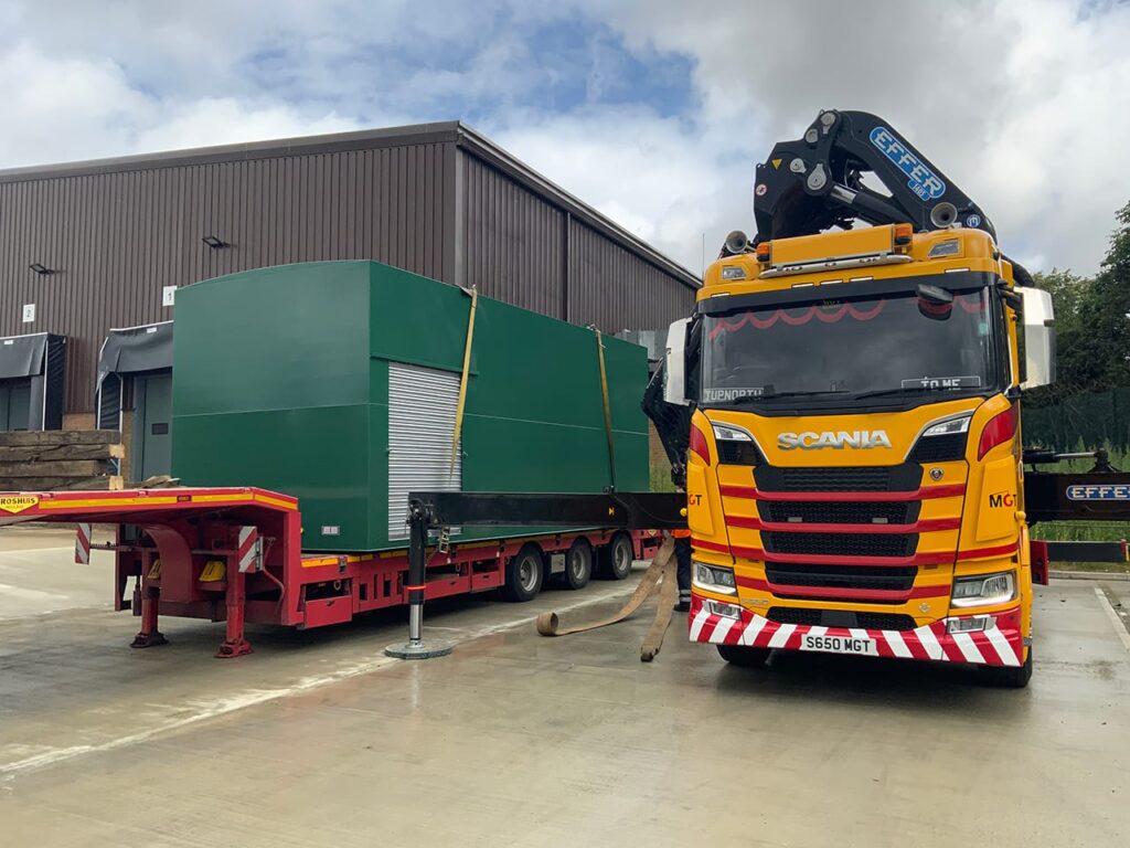 A large green Merridale commercial fuel storage tank with an integrated roller-shutter cabinet sits secured on a red flatbed trailer. A bright yellow and red Scania hiab crane truck is parked next to it, deploying its stabilization outriggers in a concrete industrial yard in front of a warehouse.