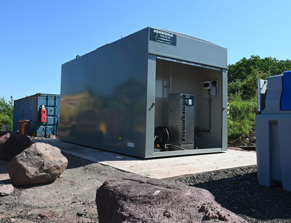 A large, grey Merridale fuel storage tank situated on a concrete pad in an outdoor industrial or construction site. The tank features a built-in secure compartment housing a stainless steel diesel dispensing pump and a heavy-duty hose reel.