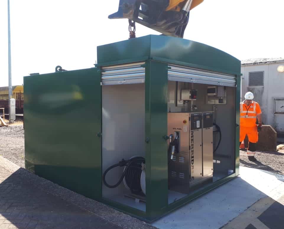A green Merridale commercial fuel storage tank being positioned on a concrete base by an overhead crane at an industrial site. The tank features an integrated walk-in cabinet with a roller shutter door, housing a dual-nozzle stainless steel fuel dispenser labelled "GTL FUEL" alongside a heavy-duty hose reel, with a site worker in high-visibility clothing looking on.