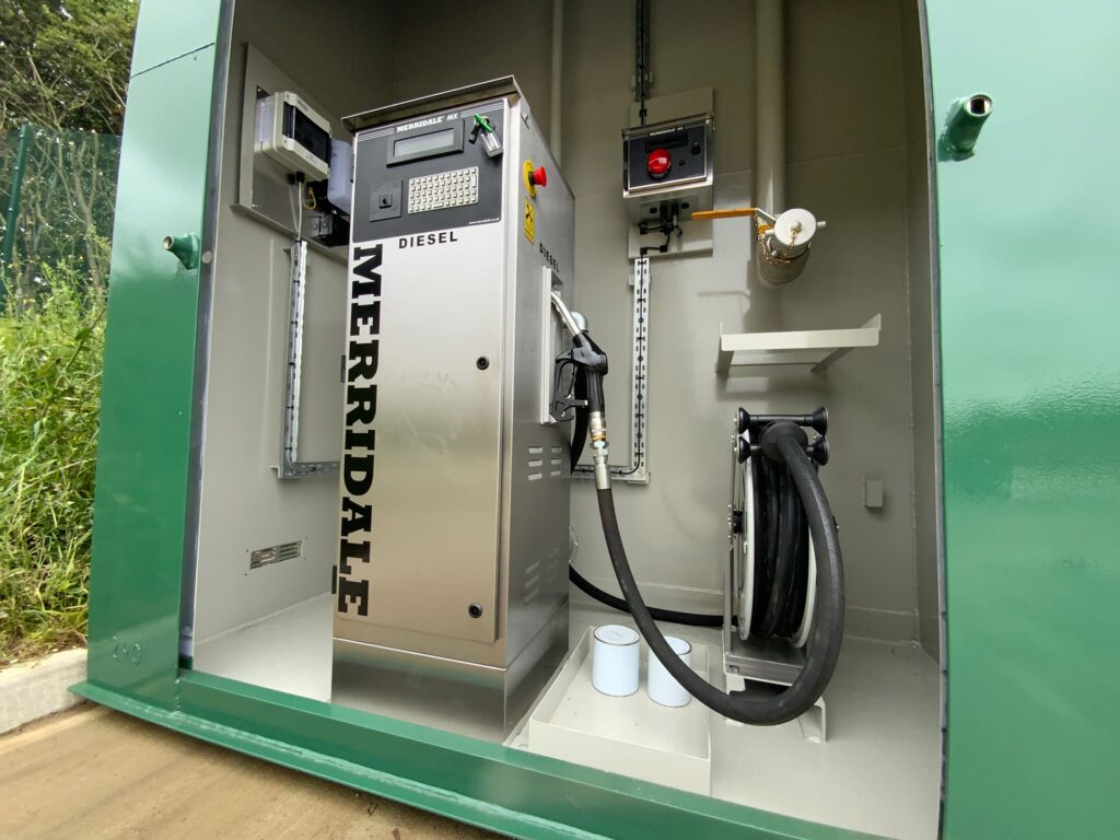 A close-up shot inside a secure green metal housing displaying a stainless steel Merridale MX diesel commercial fuel dispenser. The setup features an integrated digital management keypad, a fuel nozzle connected via a long hose to a heavy-duty storage reel, and internal electrical and piping installations.