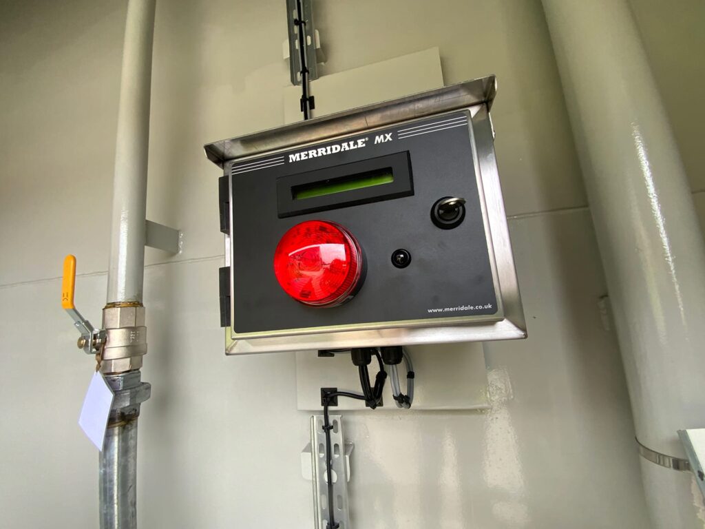 A close-up shot of a wall-mounted Merridale MX fuel management terminal box inside an industrial enclosure. The unit features a small digital display screen, a key switch, a prominent red dome warning light, and connected wiring underneath, positioned alongside commercial piping with a shut-off valve.
