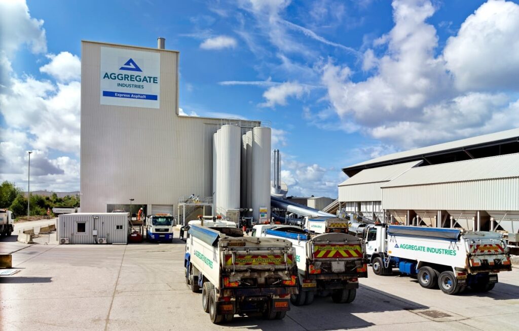 An outdoor view of an Aggregate Industries Express Asphalt plant on a sunny day with scattered clouds. Several commercial tipper trucks branded with "Aggregate Industries" and "Highway Maintenance" are parked in the concrete yard in front of large storage silos and the main processing building.