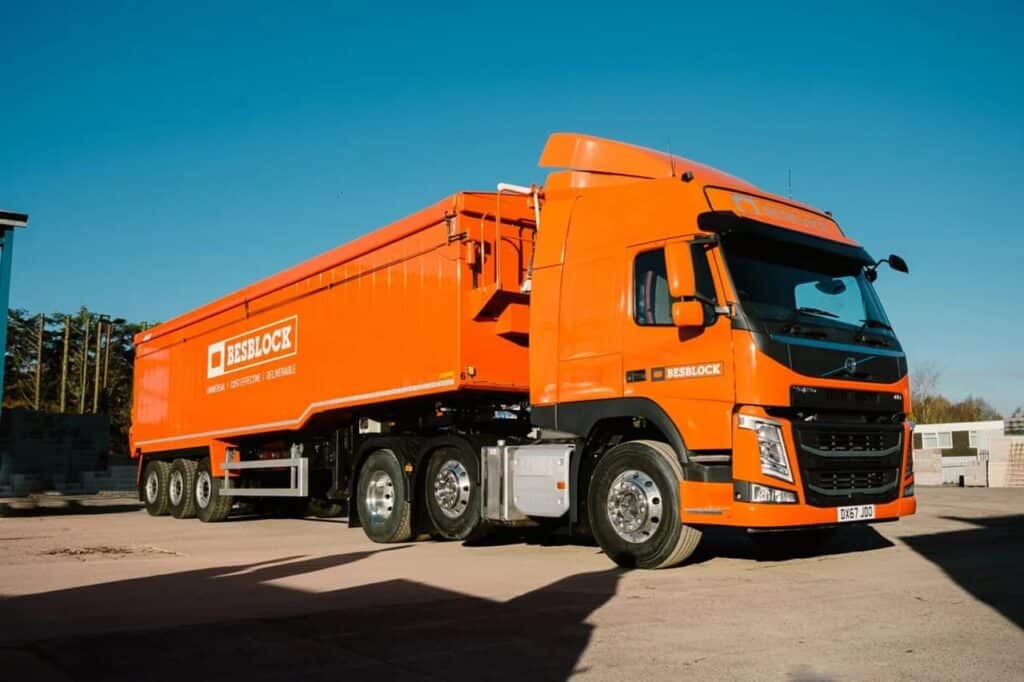 A bright orange Besblock commercial delivery truck with a large tipper trailer parked in an open concrete yard under a clear blue sky. The Volvo tractor unit features Besblock branding on the cab and trailer sides.