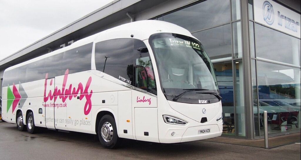 A white Linburg commercial tour coach parked outside an Irizar dealership building. The luxury coach features pink Linburg branding on the side, tinted windows, and three axles, showing an Irizar i6s model design.