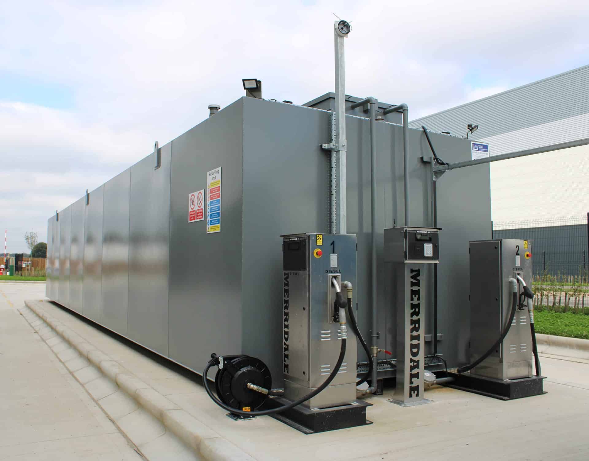 A large, industrial grey steel fuel storage tank installed on a outdoor concrete bund. Two stainless steel Merridale commercial fuel dispensing pumps and a standalone access pedestal terminal are mounted alongside the tank setup with integrated pipework and a heavy-duty hose reel.