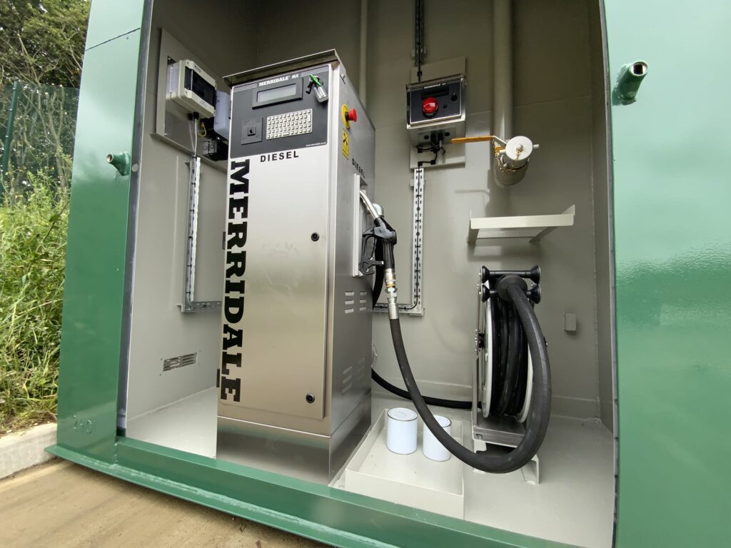 An internal close-up shot of a green walk-in industrial housing unit, displaying a high-quality stainless steel Merridale MX diesel dispenser integrated with a fuel management terminal, an internal electrical system, a shut-off valve, and a spring-loaded hose reel.